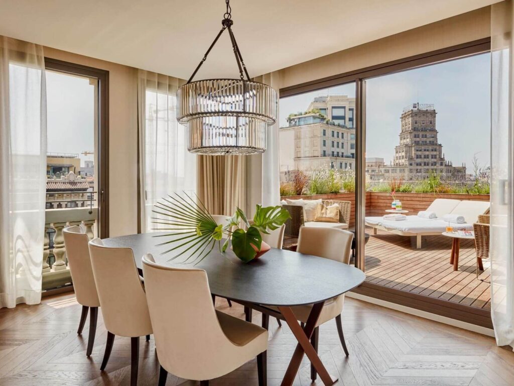 Modern dining room in a Penthouse Suite with six white chairs around a black oval table, large windows, a chandelier, and a terrace with outdoor seating overlooking city buildings.